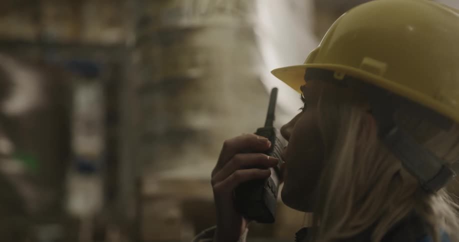 Female worker wearing yellow hard hat talking on a walkie talkie in a warehouse