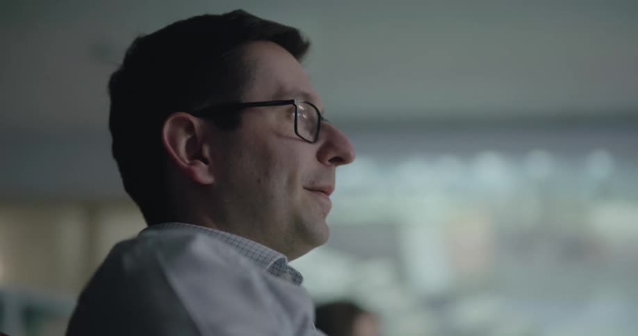 A man with glasses talks during a meeting in a large room with a projector screen