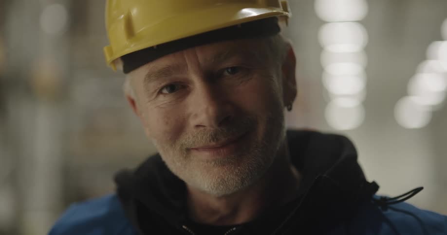 A graying warehouse worker smiles into the camera as he places a hardhat on his head