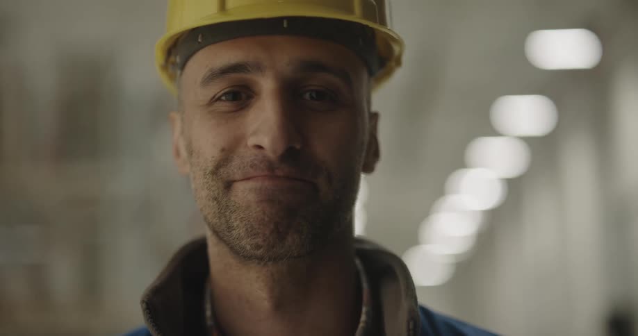 A man with a vest and a hardhat in a warehouse with lights in the background