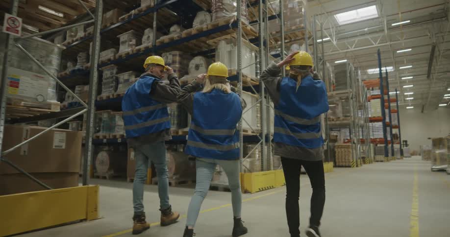 A group of workers in personal protective equipment talk with one another while walking through a warehouse before waving goodbye to one as he parts the group