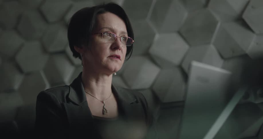 A woman with glasses and a necklace in a room with a wall in the background