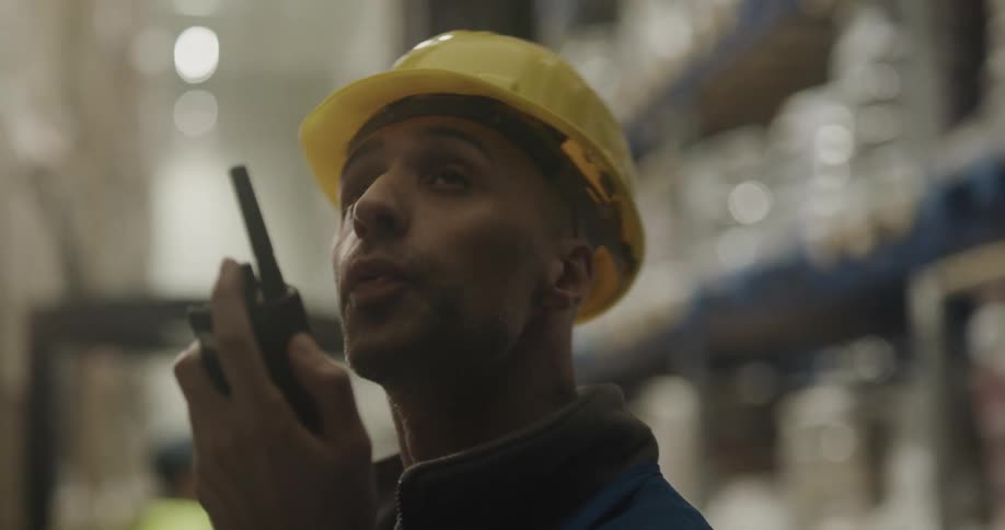 A man with a hardhat and a walkie talkie in a warehouse with shelves in the background