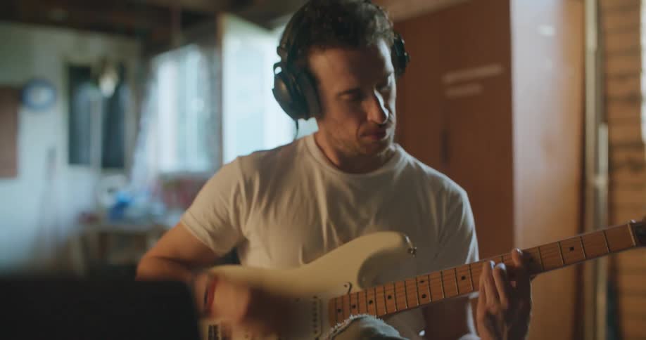 A man wearing headphones presses a button on a laptop before beginning to play the guitar while sitting on a stool indoors during the day