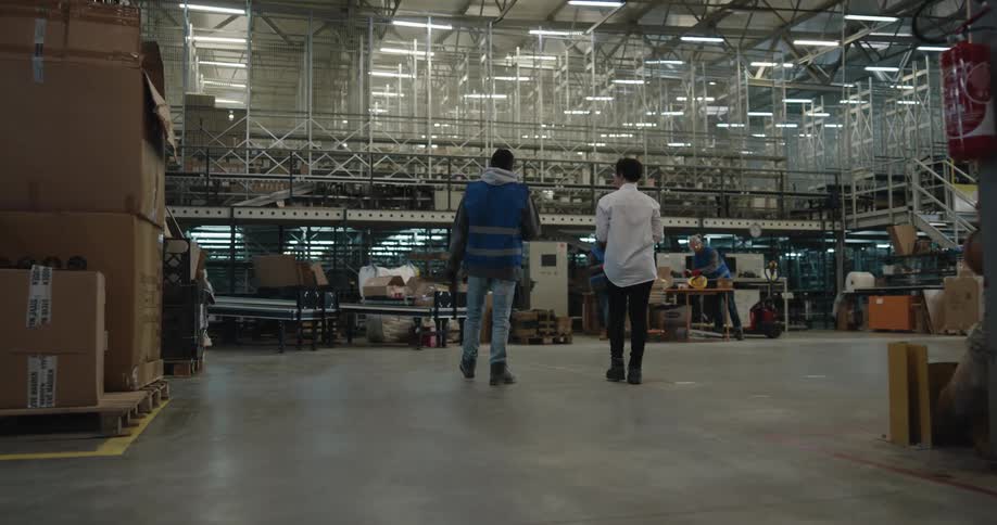 A man in a white shirt showing another man with a vest and gloves around in a warehouse with lights