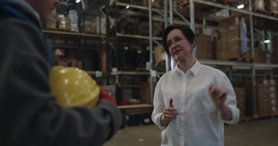 A woman with a white shirt and glasses talking to a man with a hard hat and vest in a warehouse with shelves in the background