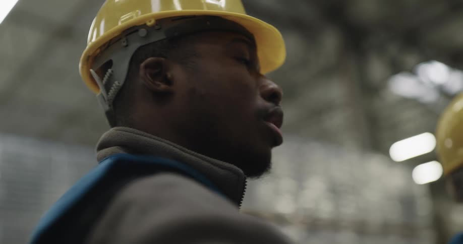 A man in a hard hat and safety vest talks to his coworker in their warehouse