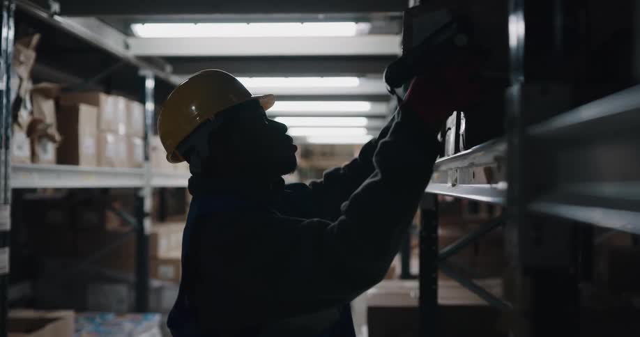 A man with a hardhat and vest scanning a box and removing it from a shelf in a warehouse