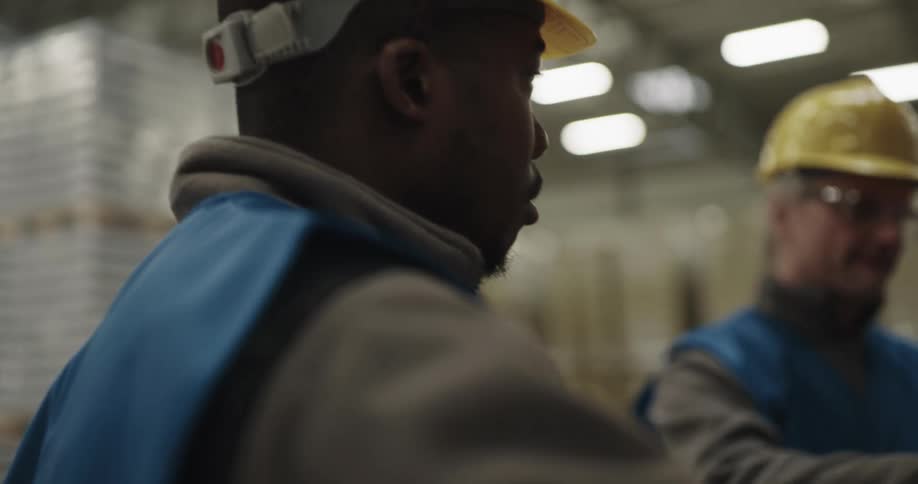A worker in a warehouse speaks into his handheld radio as an older coworker speaks to him