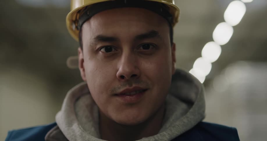 A man with a hardhat and a vest standing in a warehouse with lights in the background