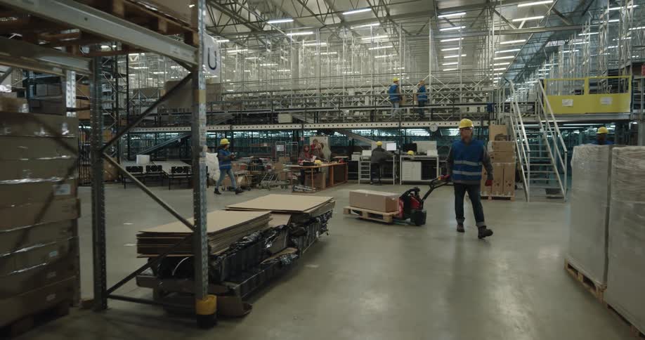 A man with a hardhat and a vest pulling a forklift with a box on a pallet in a warehouse
