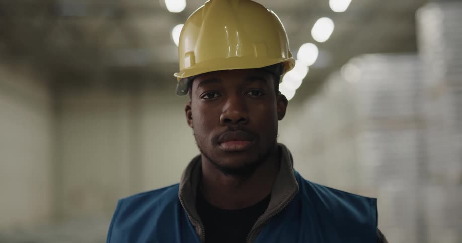A man with a vest and a hardhat in a warehouse with lights in the background