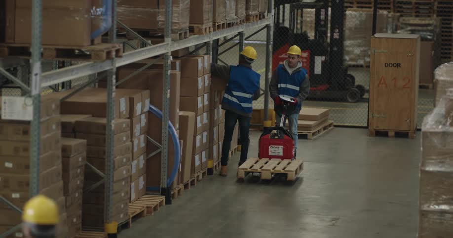 Two men in a warehouse with vests and hardhats as one scans a box and places it on a forklift