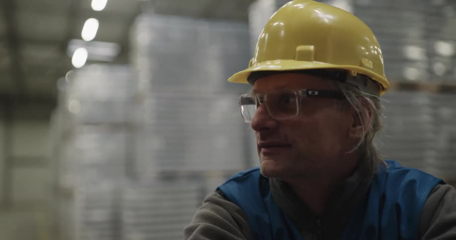 Two men in a warehouse with vests and hardhats talking with lights in the background