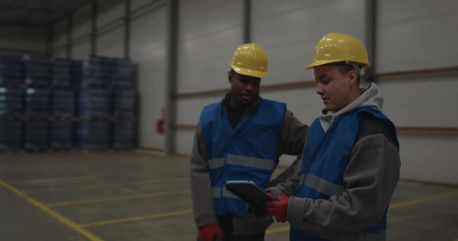 A pair of men standing in a warehouse speak with one another while looking at a tablet before walking away together