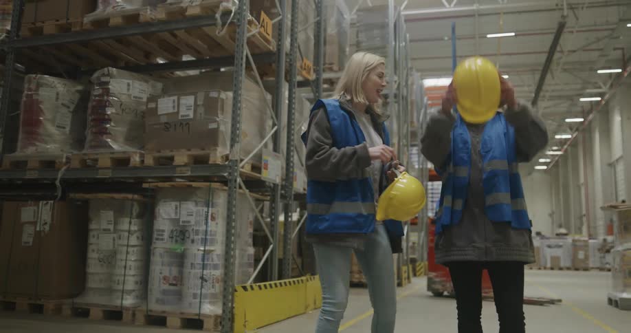 Two women walk in a warehouse talking and laughing before putting on their hardhats and going to work