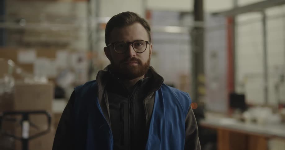 Male warehouse worker with a beard and glasses faces the camera with eye contact