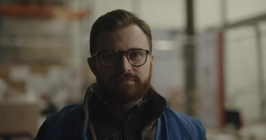 Male warehouse worker with glasses and a beard looks at the camera smilingly in the warehouse
