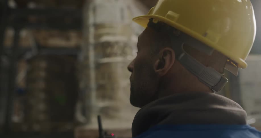 A man with a walkie talkie and a hardhat in a warehouse with shelves in the background
