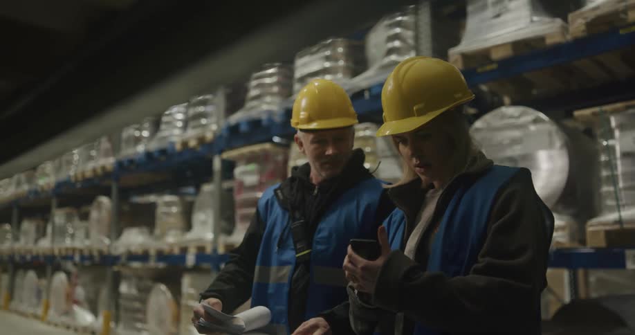 A man holding a clipboard points at a page while talking with a coworker in a warehouse