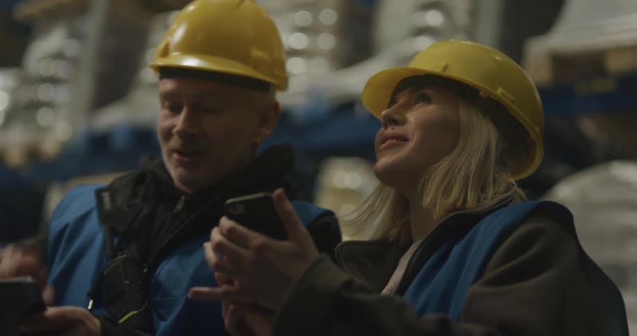 Two workers consult clipboard and mobile phone in warehouse aisle