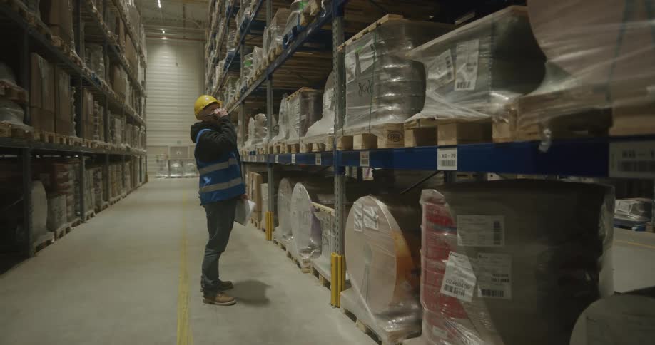 A man checks his clipboard then talks on his walkie talkie while looking up at shelves in a warehouse