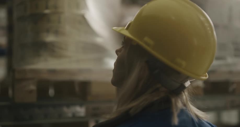A woman with a walkie talkie a hardhat and a vest in a warehouse with shelves in the background