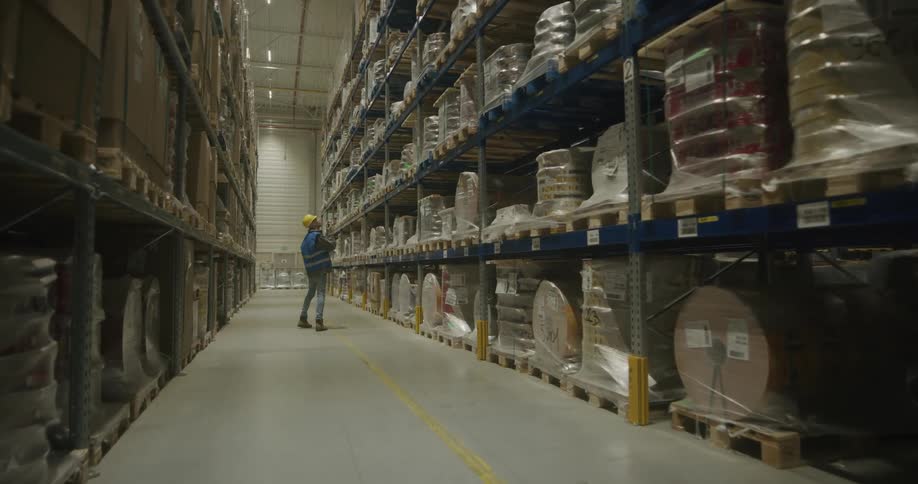 A person with a vest and a hardhat walking in a warehouse with shelves