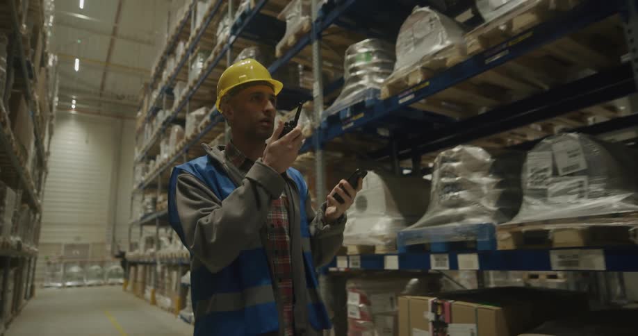 Warehouse worker walks down aisle looking at inventory while communicating via walkie talkie