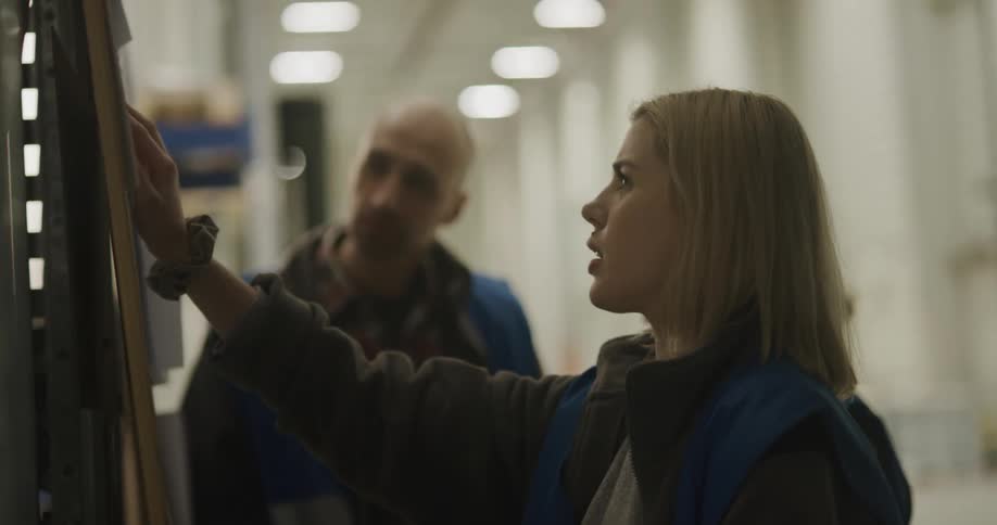 A woman with a vest in a warehouse talking to a man with a forklift and lights in the background