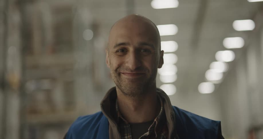 A man in a warehouse with a vest standing and smiling with shelves and lights in the background