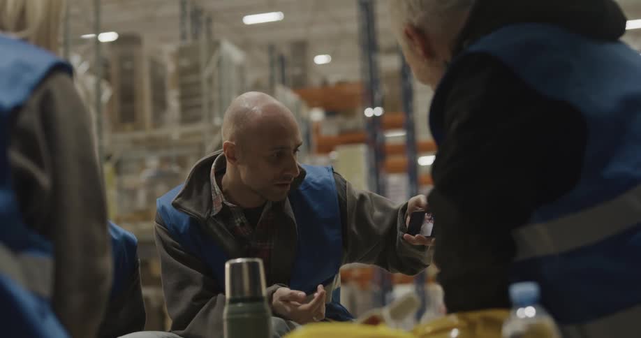 A man with a smart phone and a vest in a warehouse showing videos to a man and two women