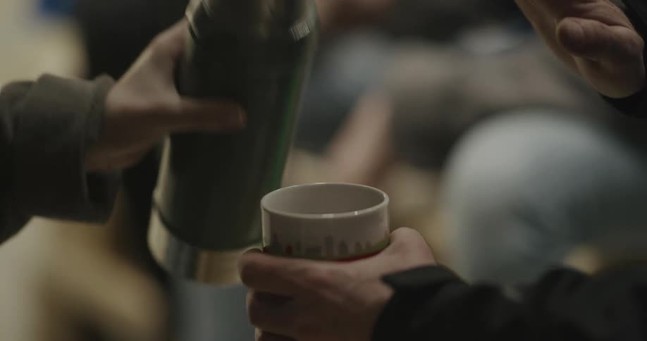 A man holds out a coffee cup as coffee is poured into it from a thermos before bringing the cup to his mouth to take a drink