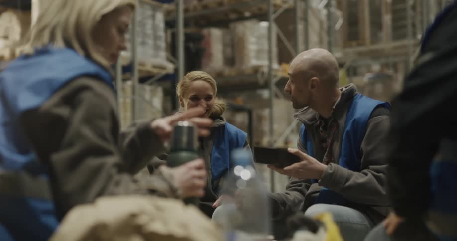 A group of warehouse coworkers sit on the ground near shelves eating and drinking during a lunch break