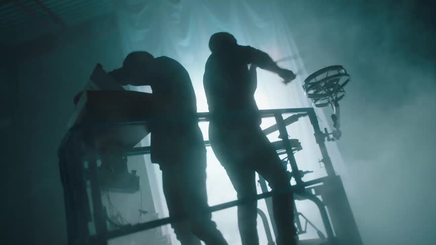 A keyboardist and a drummer play on scaffolding as smoke rises around them