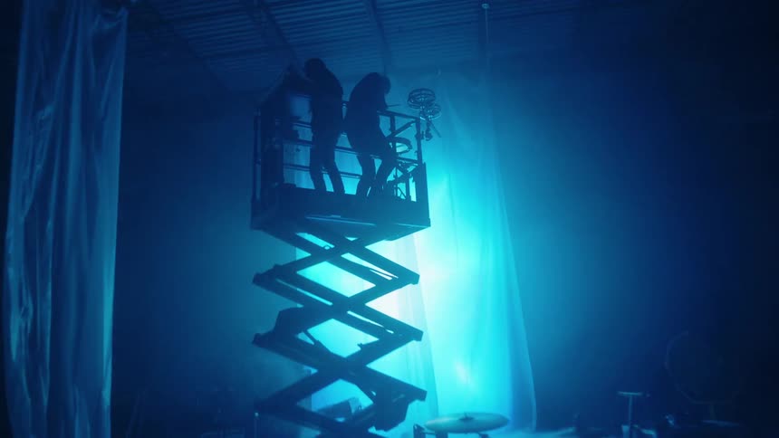 Two musicians play a keyboard and drums on scaffolding in a sound stage