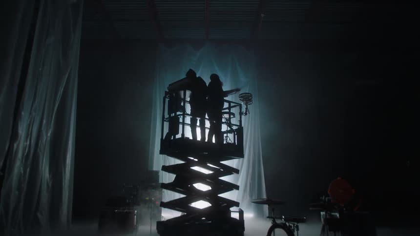 Two musicians playing instrument in a lift in front of an electronic display in the wall of a smoky dark room