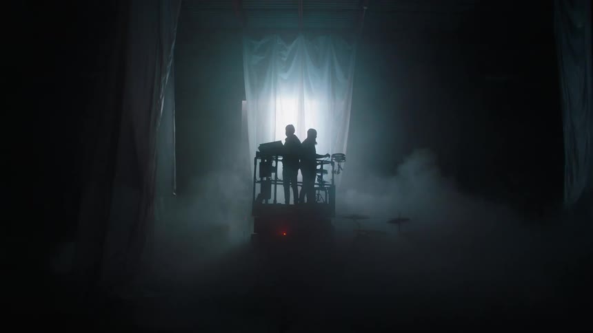 Silhouette of two men standing on a hydraulic lift as it lowers down in front of a curtain with a light strobing upwards