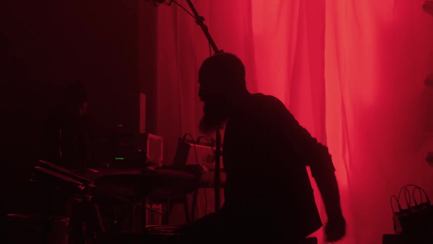 Silhouette of a man playing a drum set sitting in front of a curtain with a red light behind it
