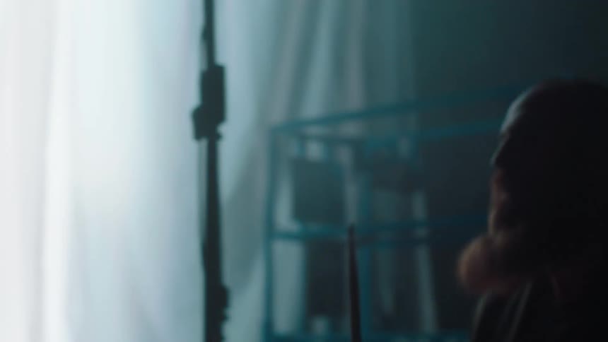 Silhouette of a man playing a drum set hard in front of a curtain with a white light flashing sporadically