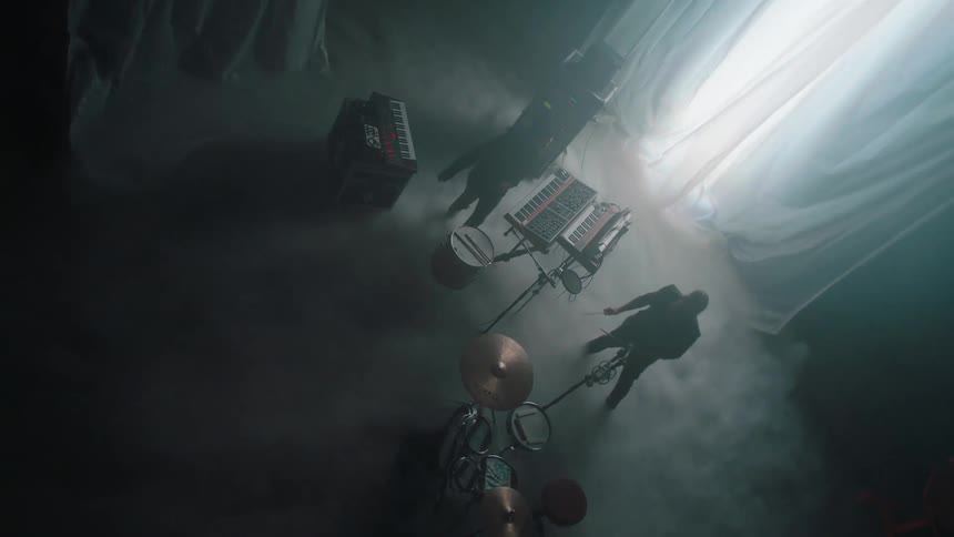 Musicians playing drums and keyboards on a stage with white smoke and flashing lights