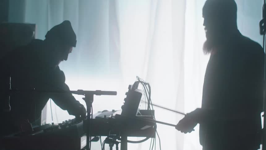 A man carrying a pair of drum sticks walking in a room with flashing white lights towards another man adjusting settings on an electronic keyboard