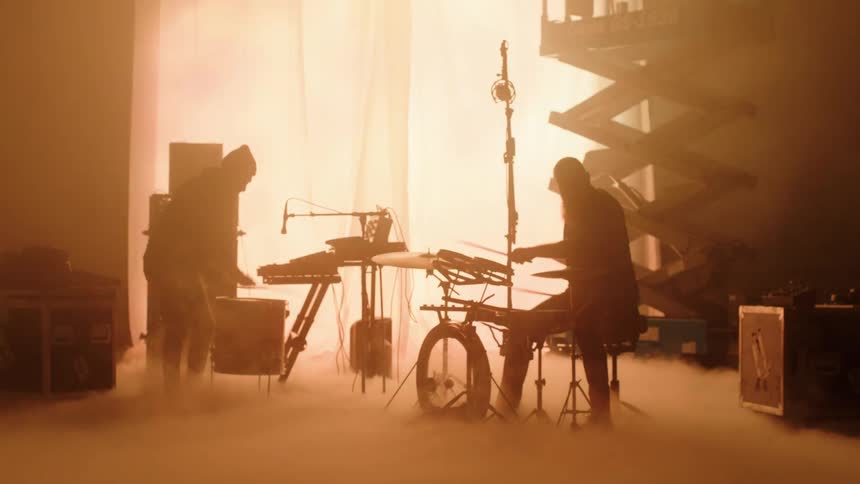 Silhouettes of two men playing a drum set on a foggy stage surrounded by stage equipment