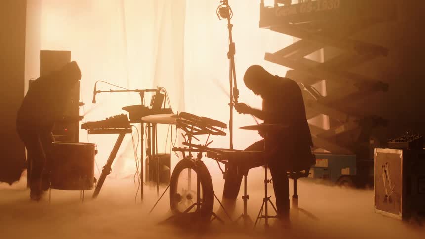 Two musicians playing drums on a smoky stage in front of a giant digital display