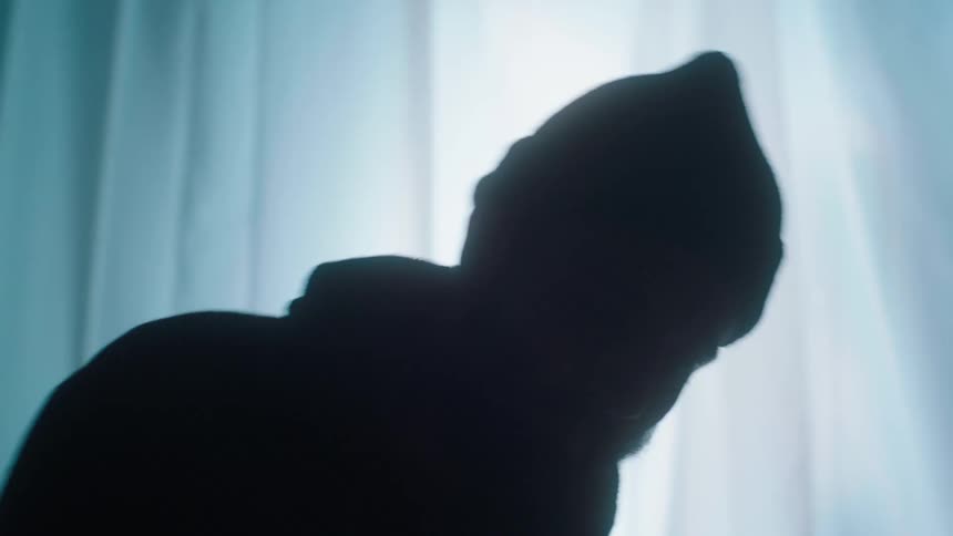 A silhouette of a man bobbing his head while he drums on a drum set in a room with a curtain and a strobing light