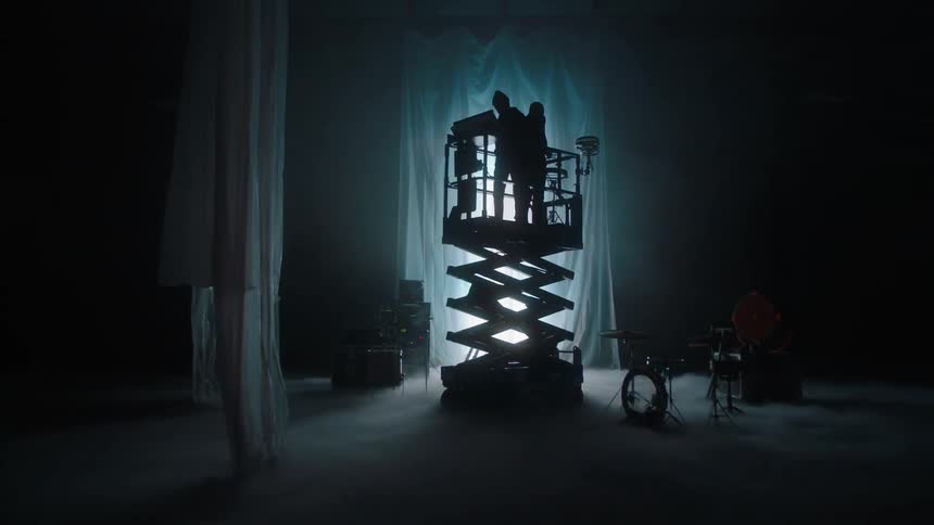 Two people on a lift in a room one playing drums and the other playing a keyboard