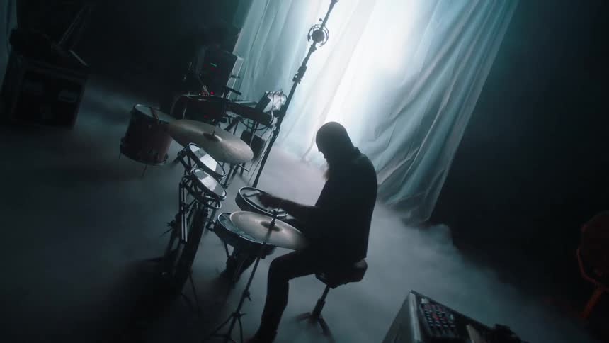 Silhouettes of two men playing musical instruments in front of a white curtain where where a white light flashes behind on a stage with smoke