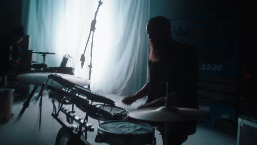 A drummer and keyboardist play on an empty stage in front of a large white curtain
