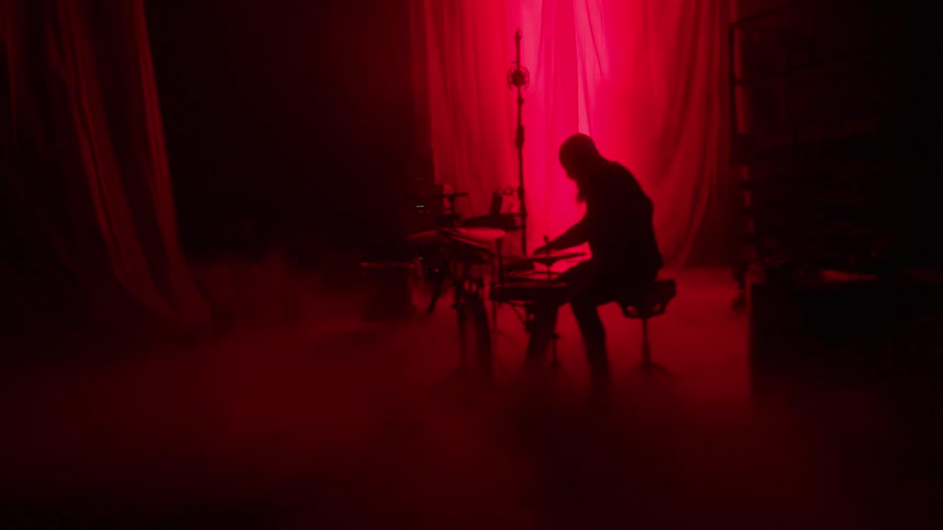 A man plays an electric piano keyboard while another man plays a drum set on stage with fog where a light is flashing behind a curtain