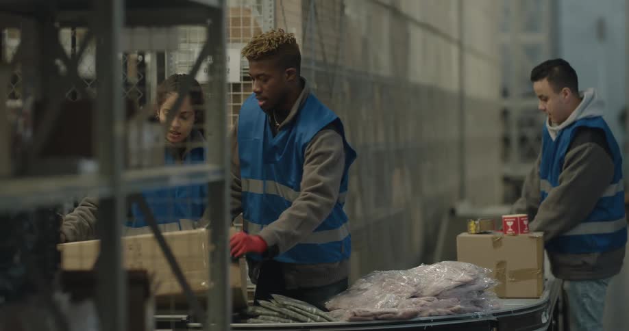 Three people wearing safety vests pack and label boxes in a warehouse